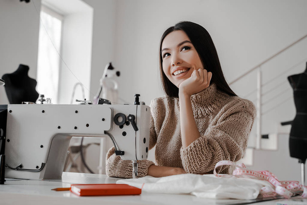 Young beautiful asian woman thinks and dreams while sitting near a sewing machine. Sewing, design work, tailoring studio, tailor, designer clothes, manufactory, in the process of creative development - Foto, imagen