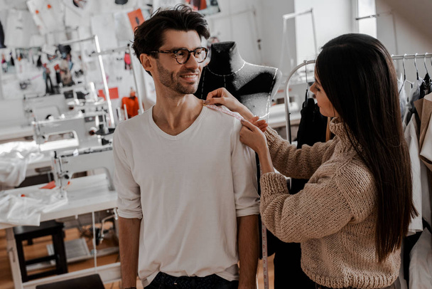 A seamstress woman takes measurements from a handsome stylish man in her sewing studio. Design, studio for sewing and cutting clothes, designer clothes, manufacturing, craft product. - Foto, Bild