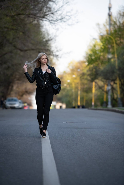 A young beautiful blonde girl in a black leather jacket is walking along an empty city road, the lower angle of the shooting - Foto, Bild