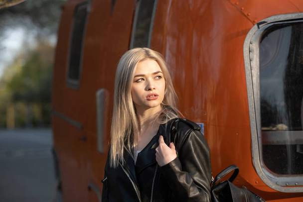 A young beautiful blonde girl in a black leather jacket stands next to an orange van - Valokuva, kuva