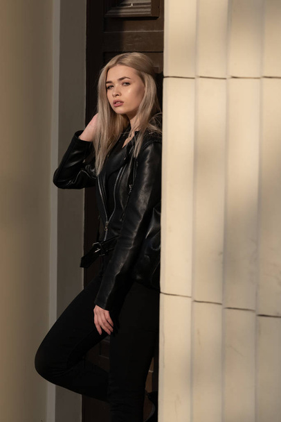 a beautiful young blonde girl stands in a black leather jacket on the background of a building - Fotó, kép