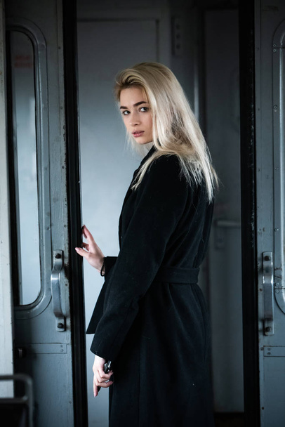 A young beautiful blonde girl in a black coat poses in the doorway of an electric train - Φωτογραφία, εικόνα