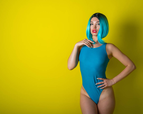 Beautiful girl in a blue wig and a bright bikini posing on a yellow background. Woman with artificial hair and a swimsuit emphasizing the buttocks. - Foto, Imagen
