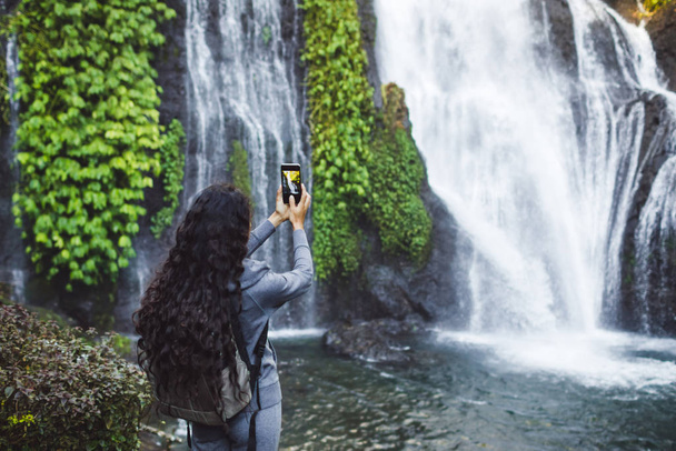 Een vrouw die een foto neemt van de waterval Banyumala in Bali. Lange krullende  - Foto, afbeelding