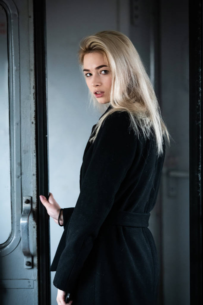 A young beautiful blonde girl in a black coat poses in the doorway of an electric train - Fotoğraf, Görsel