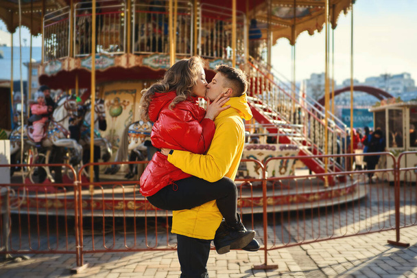 young romantic couple wearing in bright yellow and red down jackets kissing, hugging. Handsome man holding young attractive woman on french carousel background. - Foto, imagen