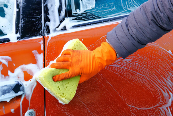 Cleaning with soap suds at self-service car wash. Man in rubber gloves washes his orange auto with yellow sponge. Soapy water runs down. - Zdjęcie, obraz