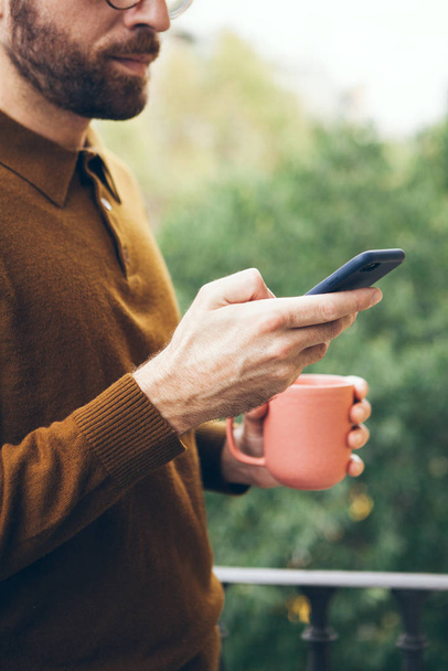 Handsom γενειάδα άνθρωπος πίνει τσάι και κρατώντας το κινητό τηλέφωνο σε μπλε περίπτωση, ενώ χρησιμοποιώντας το κοινωνικό δίκτυο / ανάγνωση ειδήσεων, σερφάροντας στο διαδίκτυο / internet στο τηλέφωνο. Επιλεκτική εστίαση. - Φωτογραφία, εικόνα