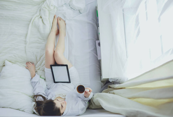 Portrait of woman using tablet and drinking coffee while sitting on the bed in the morning - Фото, зображення