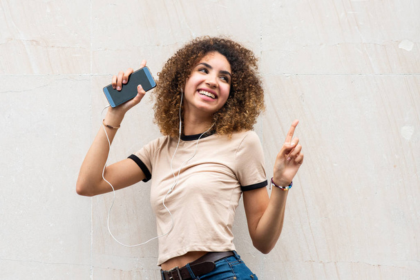 Portrait happy young african american woman dancing with mobile phone and earphones while listening to music by wall - 写真・画像