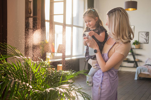 Молодая привлекательная женщина со своей маленькой девочкой поливает растения в доме. Работа по дому
 - Фото, изображение