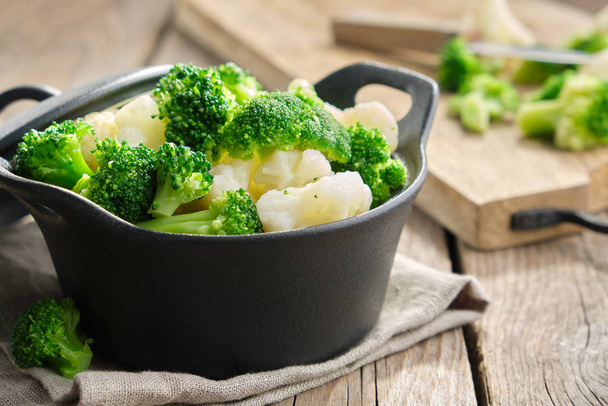 Black saucepan with cooked broccoli and cauliflower on wooden kitchen table. - Fotó, kép