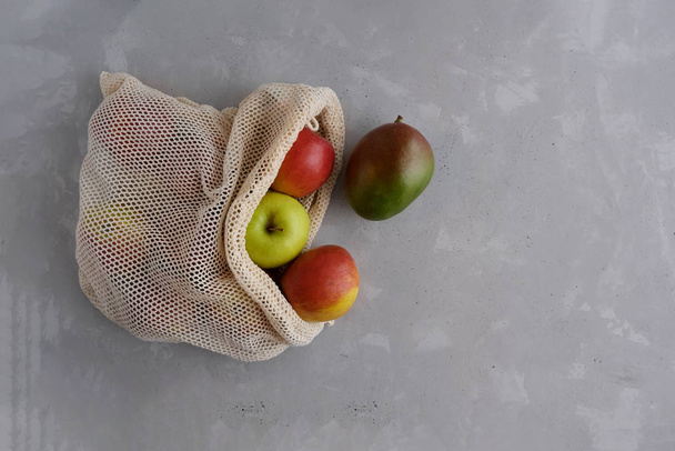 Fresh juicy red and yellow apples and mangoes with a cotton reusable beige bag bag on a gray concrete background. Fruit in a string bag. Place for text. No waste. - 写真・画像