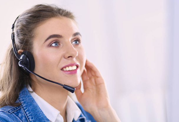 Χαρούμενη γοητευτική νεαρή γυναίκα που κάθεται και εργάζεται με φορητό υπολογιστή χρησιμοποιώντας ακουστικά στο γραφείο - Φωτογραφία, εικόνα