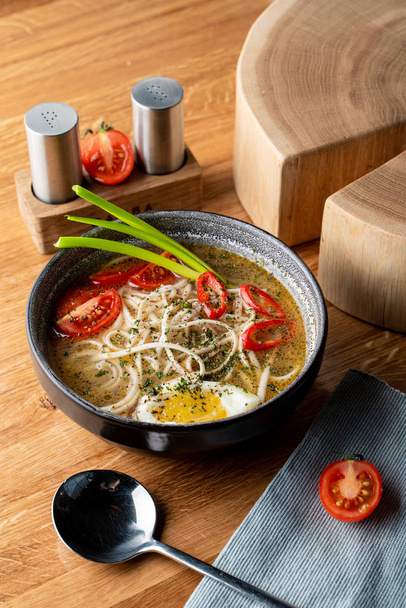 Hot chicken noodle soup with boiled egg, tomatoes and chili pepper on wooden table - 写真・画像