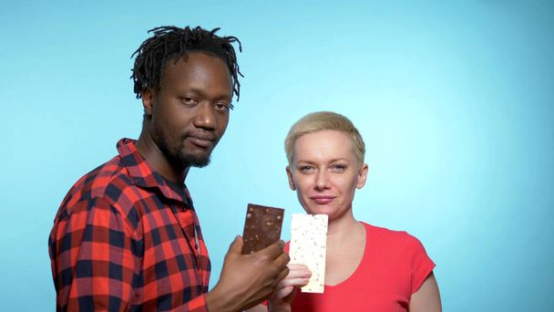 African man , Caucasian woman to eat black and white chocolate. blue background - Photo, image