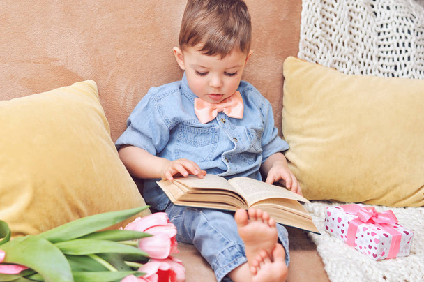 Ένα μικρό αγόρι κάθεται στο σπίτι στον καναπέ του με ένα βιβλίο για την αγάπη και τα όνειρα. - Φωτογραφία, εικόνα