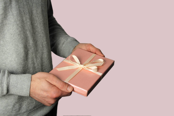 man holds a gift in his hand in a pink box with a satin ribbon on a light background, holiday concept - Foto, afbeelding
