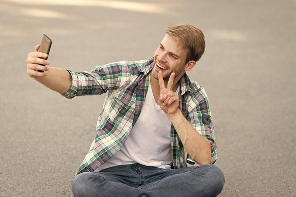 free time spending. summer fashion. happy man checkered shirt. male fashion. student relax use phone. macho man make selfie. street style. man sit on ground. carefree student. he love selfie - Foto, afbeelding