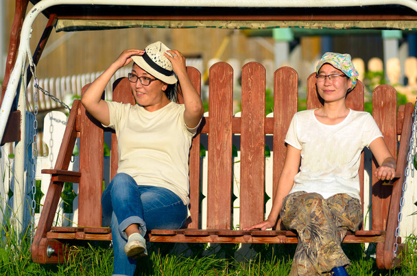 Kaksi tyttöä tyttöystäviä Aasian yakuts rentoutua keinu vieressä kukkapenkkiin asuinalueella kylässä Pohjois-Yakutia nauttia lämpimästä auringonvalosta
. - Valokuva, kuva