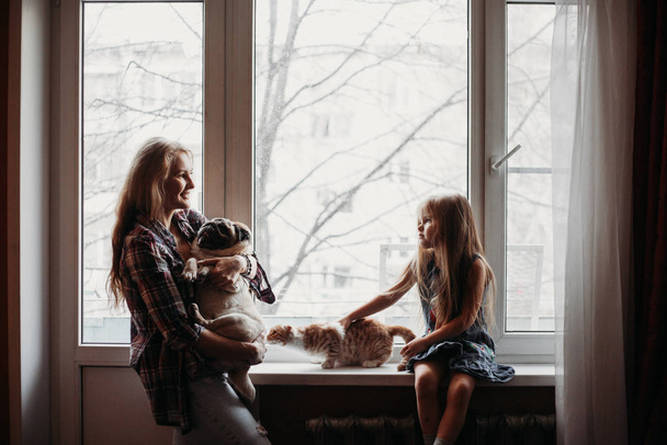 Mom holds a dog and stands near the window, the girl sits near the window, the family home - Foto, Bild