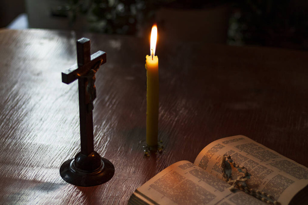 Buntglastisch aus Holz. Es gibt ein Buch darüber. Darauf steht ein Kreuz, eine Kerze brennt. schummriges Licht. Dunkelheit. Es gibt eine Tönung - Foto, Bild