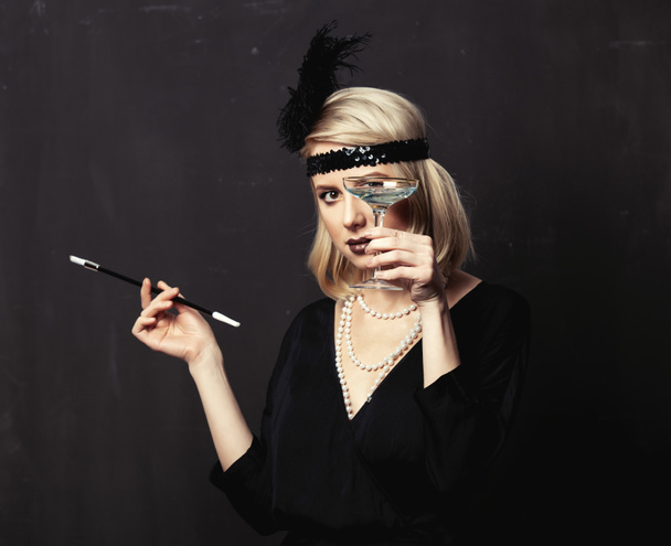Beautiful blonde woman in twenties years clothes with smoking pipe and cocktail on dark background - Fotografie, Obrázek