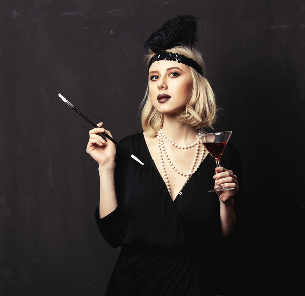 Όμορφη ξανθιά γυναίκα σε είκοσι χρόνια ρούχα με πίπα καπνίσματος και κοκτέιλ σε σκούρο φόντο - Φωτογραφία, εικόνα