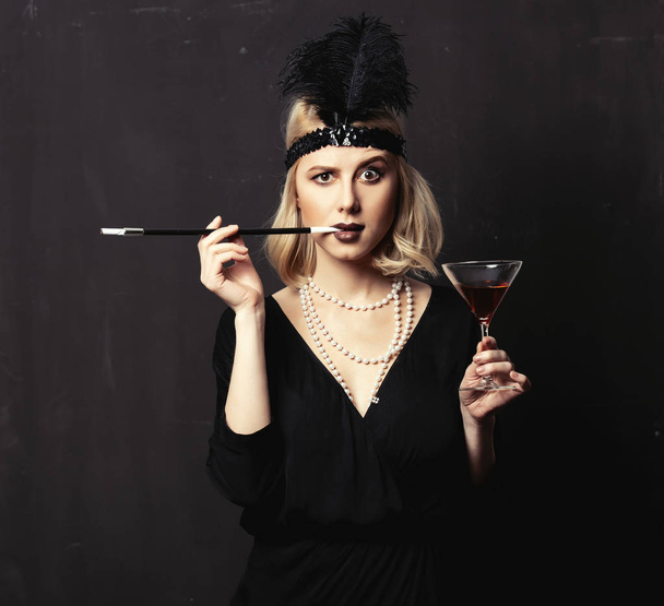Beautiful blonde woman in twenties years clothes with smoking pipe and cocktail on dark background - Photo, Image
