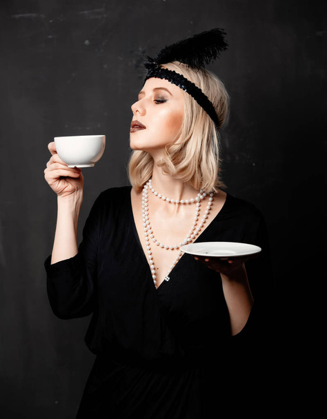 Krásná blondýna žena ve dvacátých letech oblečení s šálkem kávy na tmavém pozadí - Fotografie, Obrázek