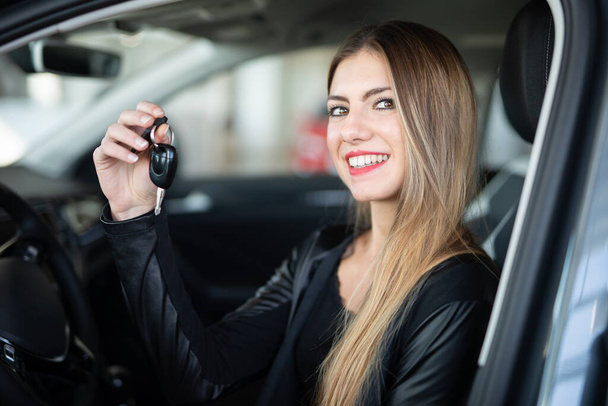 Young happy woman showing the key of her new car in a car dealer saloon - Valokuva, kuva