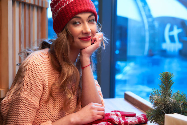 Beautiful caucasian blonde woman in red knitted hat sitting behind cafeteria window drinking a warm latte on a wonderful Christmas day. - Fotó, kép
