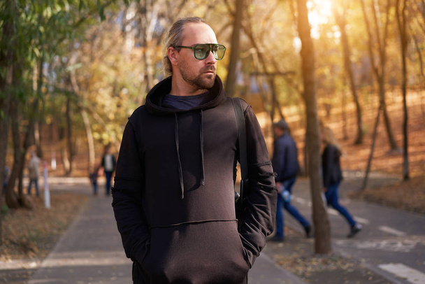 Handsome young unshaven man black hoodie and sunglasses walking autumn park warm day. - Foto, Imagen