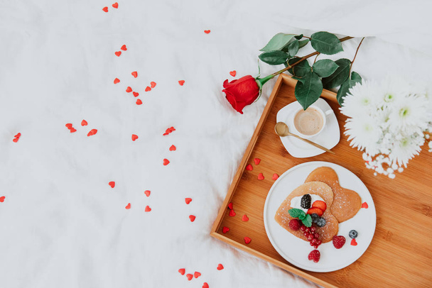 Pancakes in the shape of a heart with berries, morning  coffee and rose on white bed. Valentine's day breakfast concept - Foto, afbeelding