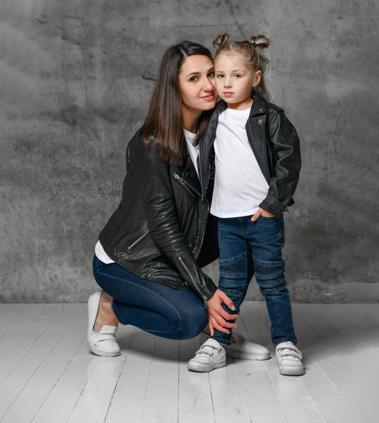Mom and child hugging each other while posing on gray studio background. Wearing in black leather jackets, white t-shirts, blue jeans. Close up - Foto, Imagen