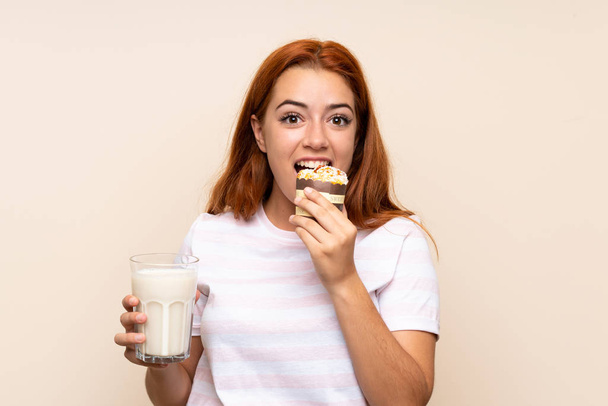 Teenager redhead girl holding a glass of milk and a muffin over isolated background - Valokuva, kuva