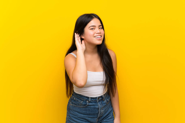 Joven adolescente asiática chica sobre aislado amarillo fondo escuchar algo poniendo la mano en la oreja
 - Foto, Imagen