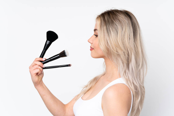 Teenager blonde Mädchen über isolierten weißen Hintergrund halten Make-up-Pinsel und schauen Sie es - Foto, Bild