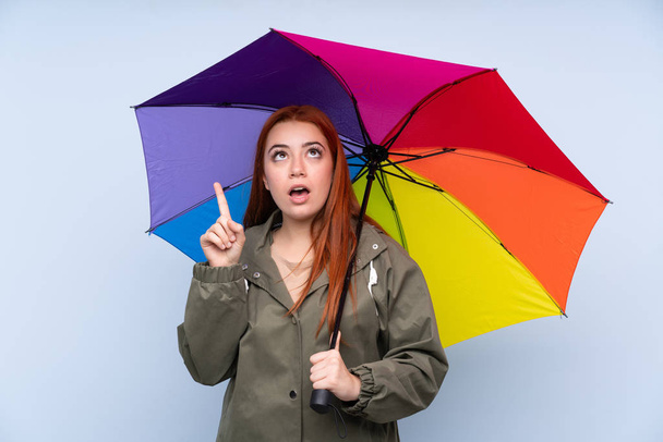 Vörös hajú tinédzser lány kezében egy esernyő felett elszigetelt kék háttér rámutatva a mutatóujj egy nagy ötlet - Fotó, kép