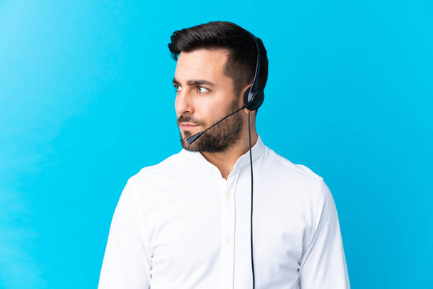 Telemarketer man working with a headset over isolated blue background looking side - 写真・画像