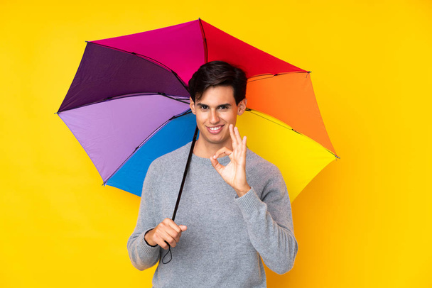 Чоловік тримає парасольку над ізольованим жовтим тлом, показуючи знак ОК пальцями
 - Фото, зображення