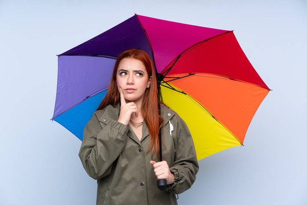 Μια κοκκινομάλλα έφηβη που κρατάει ομπρέλα πάνω από απομονωμένο μπλε φόντο και σκέφτεται μια ιδέα. - Φωτογραφία, εικόνα