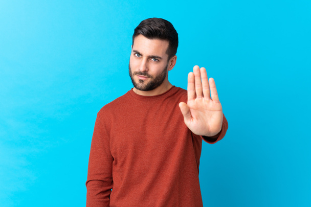 Mladý pohledný muž s vousem přes izolované modré pozadí dělat stop gesto s její rukou - Fotografie, Obrázek