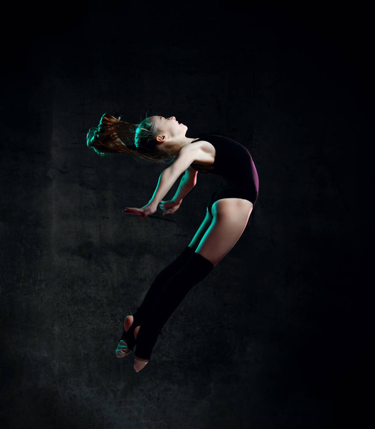 Young girl gymnast in black sport body and special uppers jumping in gymnastic pose over dark background - 写真・画像
