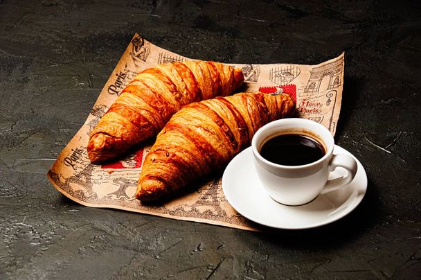 Sweet tasty croissants, white cup of strong ristretto coffee on a saucer on craft paper on a dark background with copy space - Foto, afbeelding