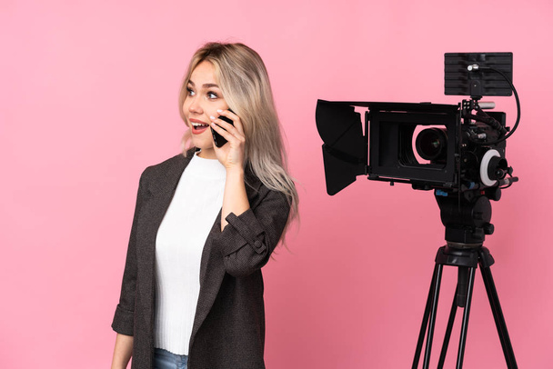 Reporter femme tenant un microphone et rapportant des nouvelles sur fond rose isolé garder une conversation avec le téléphone portable avec quelqu'un - Photo, image