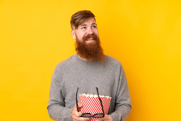 Roodharige man met lange baard over geïsoleerde gele achtergrond met een grote emmer popcorn - Foto, afbeelding