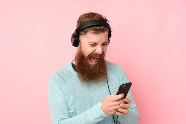 Redhead man with long beard over isolated pink background listening music with a mobile and singing - Fotografie, Obrázek
