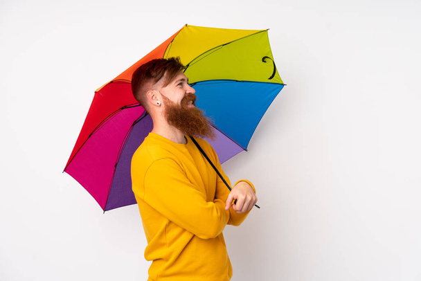 Hombre pelirrojo con barba larga sosteniendo un paraguas sobre un fondo blanco aislado mirando hacia un lado - Foto, Imagen