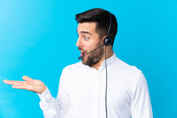 Hombre de telemarketer trabajando con un auricular sobre un fondo azul aislado sosteniendo un espacio de copia imaginario en la palma
 - Foto, Imagen